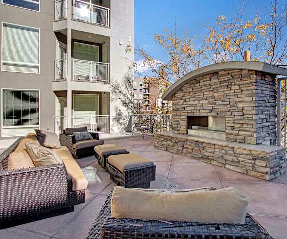 The outdoor resident lounge with plush couches and chairs with a large outdoor fireplace with a tree behind it and the gray apartment building in the background at Velo on the Boulevard Apartment Homes. 