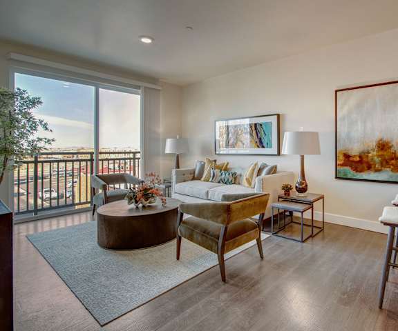 The spacious living room with a white couch, silver and gold throw pillows, large windows, a ceiling fan, beautiful wall art, plush chairs, coffee table, decorations, large windows, a TV, and glass doors to the balcony at the Ritz Classic Apartment Homes. 