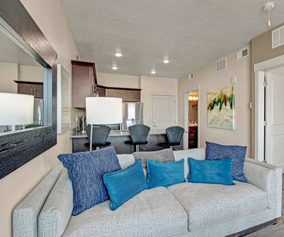 A view of the living room with a plush white couch with blue pillows, lamps, and the large kitchen with stainless steel appliances, marble countertops, and spacious wooden cabinets at Velo on the Boulevard Apartment Homes. 