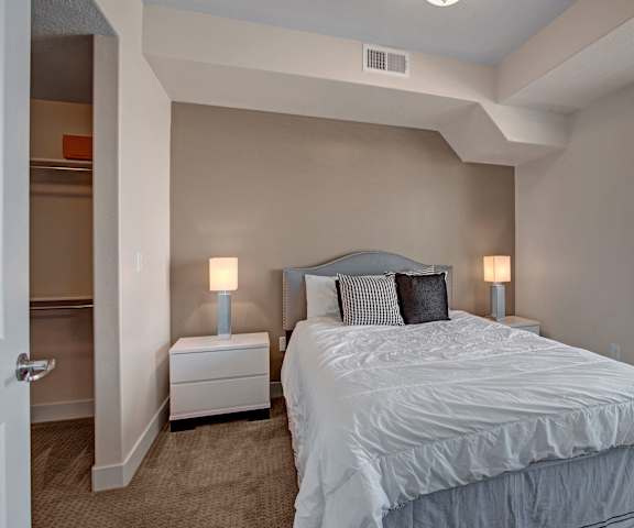 A spacious bedroom with a queen-size bed with a white and gray bedspread, a dresser, wall art, lamps, decorations, window, a closet, and nightstands at Velo on the Boulevard Apartment Homes. 