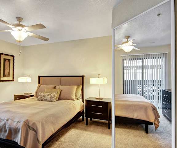 A cozy bedroom with a queen-size bed, a dresser, a mirror, a ceiling fan, decorations, window, and nightstands at Reserve at Arrowhead Apartment Homes. 