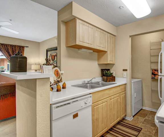 A cozy kitchen with white appliances, kitchen utensils, overhead lighting, light countertops with lots of room, a bar counter with barstools, and large wooden cabinets at Retreat at the Speedway Apartment Homes. 