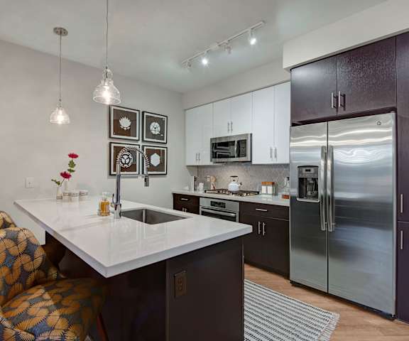 A large, modern kitchen with stainless steel appliances, kitchen utensils, white countertops with lots of room, a kitchen island with barstools and two places set for a meal, wall art, and large dark wooden cabinets at Roosevelt Row Apartment Homes. 