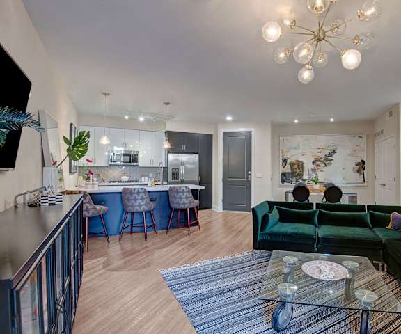 The spacious living room with a green velvet couch, a blue rug, glass coffee table, unique overhead lighting, decorations, a TV, and view of the kitchen with a bar with three barstools at Roosevelt Row Apartment Homes. 