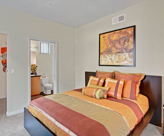 A spacious bedroom with a queen-size bed with a red and orange bedspread, a dresser, decorations, closet, and nightstands at Visions Apartment Homes. 
