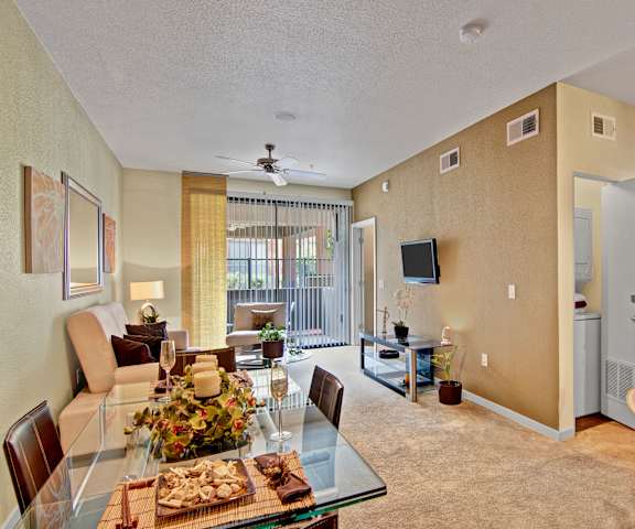 The spacious living room with a couch, plush chairs, coffee table, overhead lighting with a ceiling fan, decorations, a TV, and view of the dining room with a table, chairs, and place settings for four with champagne glasses and a plate with snacks, and the kitchen at Visions Apartment Homes. 