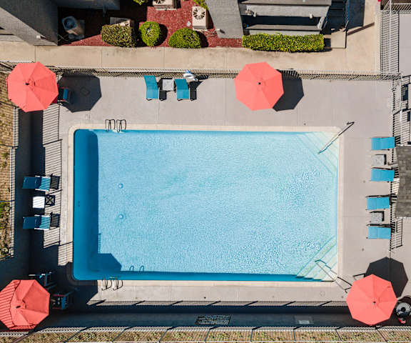 The aerial view of turquoise outdoor pool with orange umbrellas, blue pool chairs, tables, and chairs with the apartments in the background at Hillside Village Apartments. 