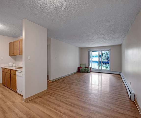 An empty living room with hardwood flooring and view of the kitchen with a door to a balcony at Gateway Gardens. 