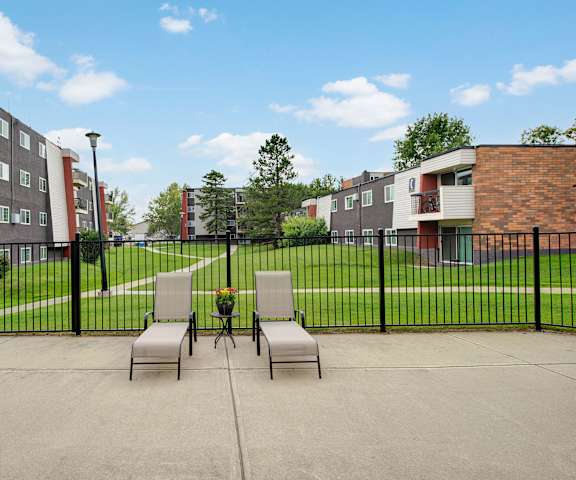 Two lounge chairs with a plant in between them in front of the green space, walkways, and apartment buildings Park Place South Apartments. 