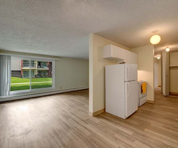 A view of the empty carpeted living room with a window and the kitchen with white appliances and wooden large cabinets at Park Place South Apartments. 