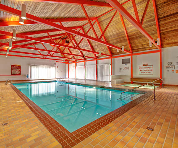The turquoise indoor pool with tables, chairs, and a jacuzzi in a large room with wooden ceiling with orange beams, light brick floors, and windows with curtains at Ranchlands Apartments Homes. 