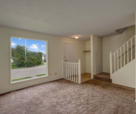 An empty living room with carpeting, stairs to the upper floor, a large window, and the front door at Ranchlands Apartment Homes. 