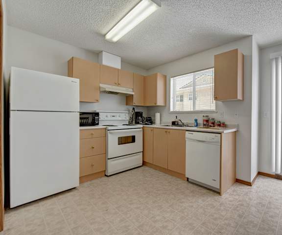 A spacious kitchen with white appliances, light counter, and large wooden cabinets with a window and sliding glass doors to the balcony at Ridgeview Village. 