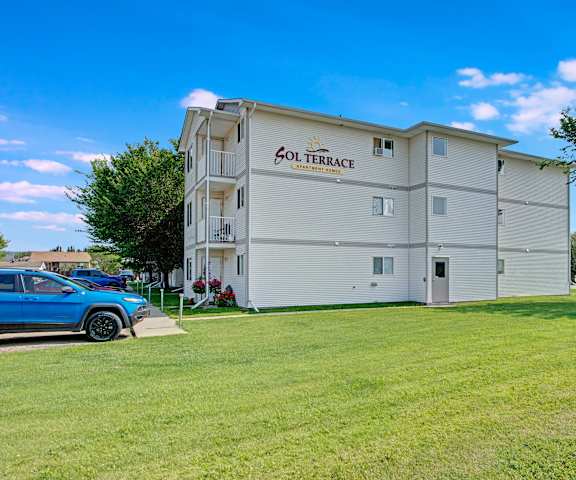 The exterior of the white Sol Terrace Apartment Homes building with the parking lot in front and surrounded by the grassy area with trees around. 