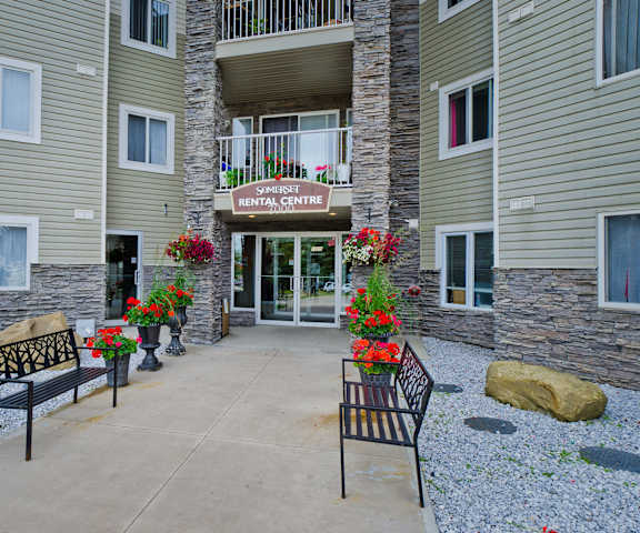 The front walkway to the rental center with a sign above the glass doors and red potted flowers lining the walkway with two benches on either side at Somerset Pointe Apartment Homes. 