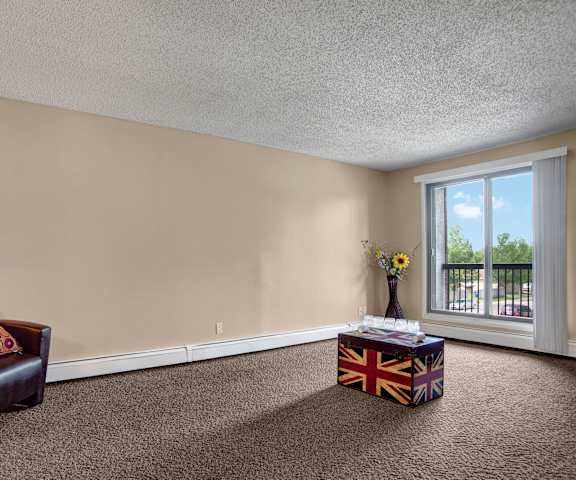 A large, empty living room with carpeting, a leather chair with orange pillows, a union jack coffee table, sunflowers in a vase, and a door to the balcony at Ventana Apartment Homes. 
