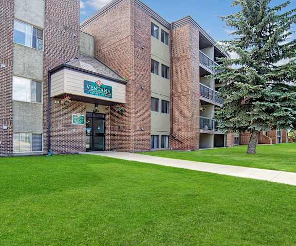 The exterior of the red brick building with the leasing office with an emerald green sign above the doors surrounded by grass with a walkway and a evergreen tree at Ventana Apartment Homes. 