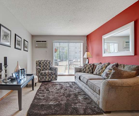 The spacious living room with a couch, throw pillows, wall art, plush chairs, coffee table, decorations, a mirror, a red accent wall, a TV, and glass doors to the balcony at Windsor Terrace Apartment Homes. 