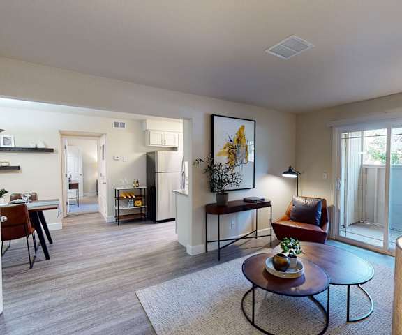 The spacious living room with a couch, plush chairs, coffee table, overhead lighting with a ceiling fan, decorations, a TV, doors to the balcony, and view of the dining room with a table, chairs, and place settings for four, and the kitchen at Retreat at Austin Bluffs Apartment Homes. 