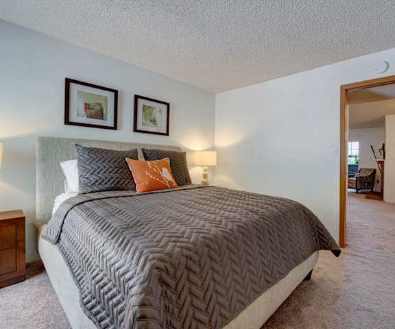 A spacious bedroom with a queen-size bed with gray bedding, a dresser, decorations, closet, and nightstands at Windtree Apartment Homes. 