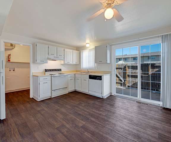 A large kitchen with white appliances, light counter, and spacious wooden cabinets with overhead lighting and sliding glass doors to the outdoor patio at Yorkshire Square Apartment Homes. 