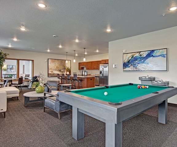 A wonderful resident lounge with tables, chairs, the community kitchen, comfortable chairs, a coffee table, and a pool table at the Club at Heritage Apartment Homes. 