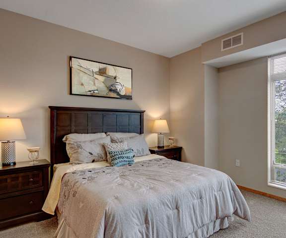 A spacious bedroom with a queen-size bed with a lavender bedspread, a dresser, wall art, lamps, decorations, large window, and nightstands with lamps at the Covington Apartment Homes. 