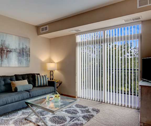 The spacious living room with a black couch, striped throw pillows, large windows, wall art, plush chair, glass coffee table, decorations, a TV, and glass doors to the balcony at the Covington Apartment Homes. 