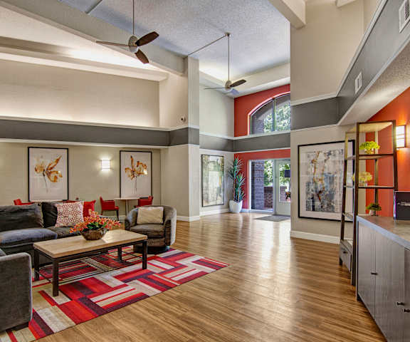 The amazing resident lounge with a gray couch, a large orange accent wall, decorations, large windows, a large TV, decorative plants, unique overhead lighting, with a colorful red and white rug, front entrance, and a coffee table at Fairways Apartment Homes. 