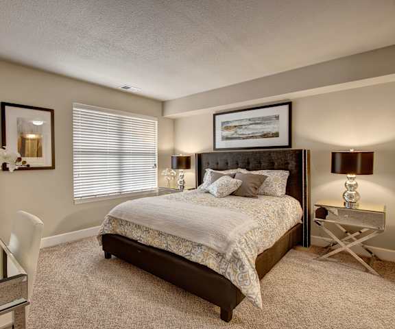 A spacious bedroom with a queen-size bed with a white and gray bedspread, a dresser, wall art, lamps, orchids, decorations, window, and nightstands with lamps at Fairways Apartment Homes. 