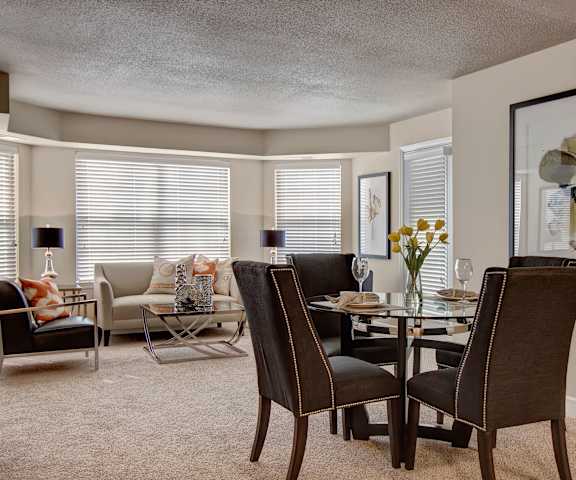 The dining room with a dining table and chairs with four places set and a view of the living room with four windows, a tan couch, and black chairs at Fairways Apartment Homes. 