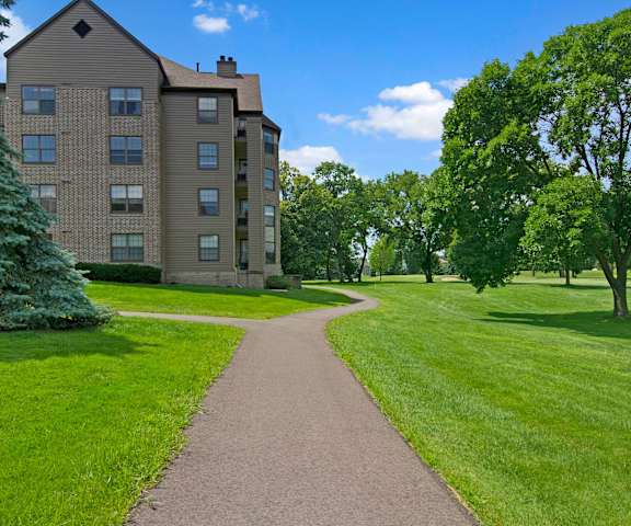 The stunning pathways surrounded by grass and trees leading to the tan brick apartment building at Fairways Apartment Homes. 