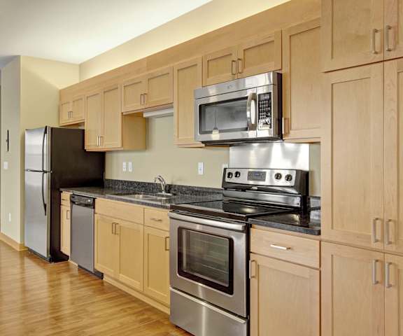 A large kitchen with stainless steel appliances, marble countertop with ample space, and large wooden cabinets at Lofts at Farmers Market Apartments. 
