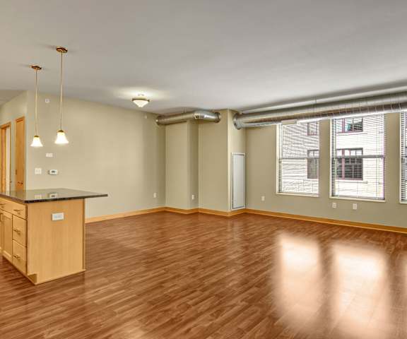 An empty living room with hardwood flooring with windows and a view of the kitchen island at Lofts at Farmers Market Apartments. 