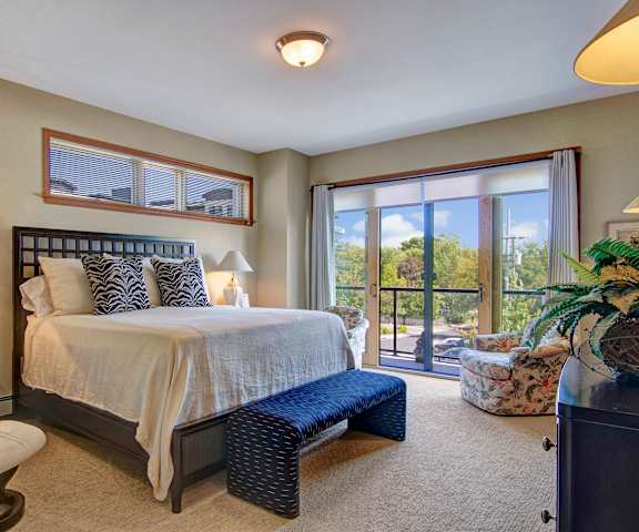 A spacious bedroom with a queen-size bed with a white bedspread, a dresser, glass doors to the balcony, a white plush chair, wall art, decorations, closet, and nightstands with lamps at the Mist at Lake Minnetonka Apartment Homes. 