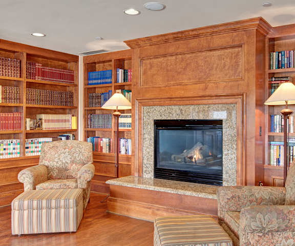 The cozy resident lounge with two floral plush chairs, two lamps, a roaring fireplace, and a beautiful library at Stoneleigh at the Reserve Apartment Homes. 