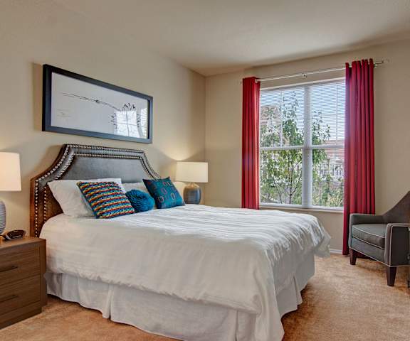 A spacious bedroom with a queen-size bed with a white bedspread, a dresser, wall art, lamps, plush brown corner chair, decorations, large window with red curtains, and nightstands with lamps at Stoneleigh at the Reserve Apartment Homes. 