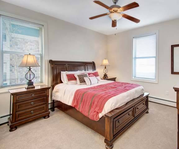 A large bedroom with a queen-size bed with white and salmon-colored bedding, a dresser, decorations, windows, and nightstands at Prairie Pines at the Ridge Apartment Homes. 