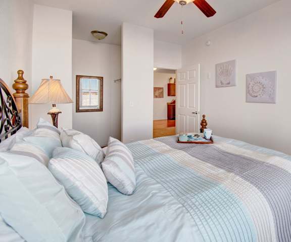 A large bedroom with a queen-size bed with light blue bedding, a small window, a dresser, decorations, closet, and nightstands at Prairie Pines at the Ridge Apartments. 