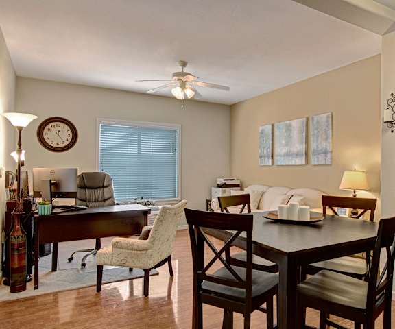 A dining room with dark wood tables and chairs, a small view of the kitchen showing the coffee maker and dishwasher, and the open living room with an office space, couch, wall art, a coffee table, a window, and a ceiling fan at Prairie Vista Apartment Homes. 