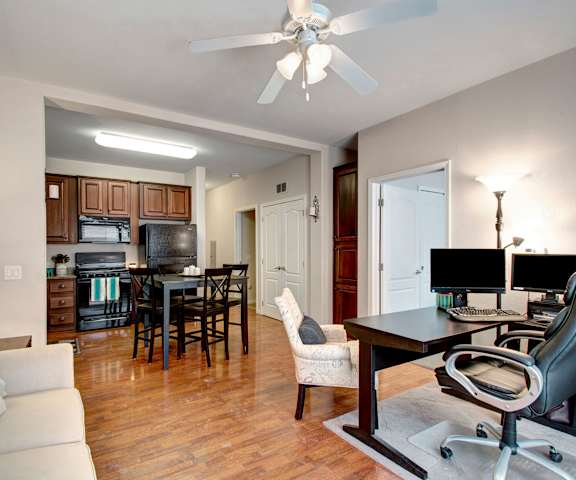 A view of the kitchen with black appliances and dark wooden cabinets and dining room with black wooden chairs and a tablefrom the living room and office with a couch and desk with an office chair and computer monitor at Prairie Vista Apartment Homes. 