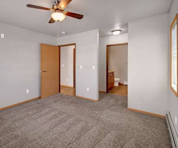 A large, empty carpeted bedroom with overhead lighting with a ceiling fan, a window, a closet, and a bathroom at Roosevelt East Apartment Homes. 