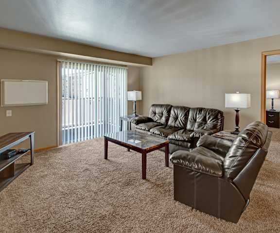 The spacious living room with a couch, throw pillows, large windows, a ceiling fan, wall art, plush chairs, coffee table, decorations, a TV, and glass doors to the balcony at Vue 28 Apartment Homes. 