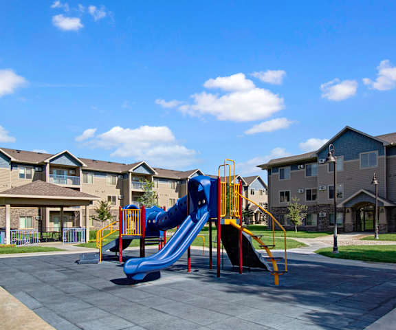 A colorful playground at a park surrounded by grass with a gazebo in the background as well as the apartment buildings at Williston Garden Apartment Homes. 