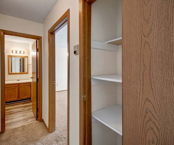 A hallway with multiple doors going to multiple rooms including a hall closet, empty bedroom, and bathroom with mirror and lighting at Windscape Apartment Homes. 