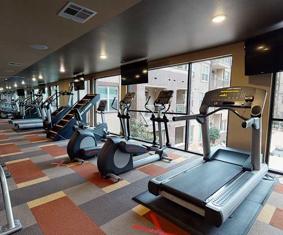 A spacious gym with cardio machines, large windows, TVs, and weights at the Edge at Midtown Apartment Homes. 