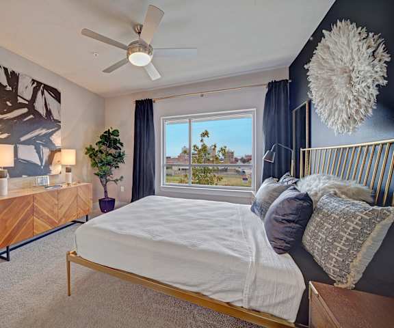 A bedroom with a queen-size bed, a dresser, decorations, wall art, window showing the surrounding buildings, and nightstands at Metropolitan Apartments. 