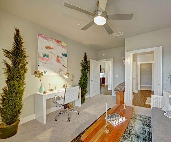 Office with a white desk and chair, coffee table with candles and a chess board, gray couch and pillows, ceiling fan, plants, decorations, wall art, and doors to the hallway and bathroom at Metropolitan Apartments. 