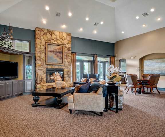 A large, well-lit resident lounge with wooden accents, windows, a door to the outside, decorations, a plush couch, bookshelves, tables, chairs, a TV, and a fireplace at Quail Landing Apartment Homes. 