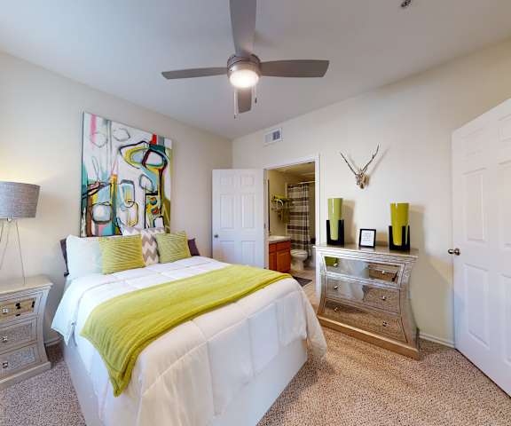 A large bedroom with a queen-size bed, a dresser, decorations, a view of the en suite bathroom, window, and nightstands at Quail Landing Apartment Homes. 